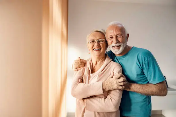 Photo of Senior couple hugging in a nursing home. A happy senior couple standing next to a window in a nursing home, hugging and smiling. They have all care they need.