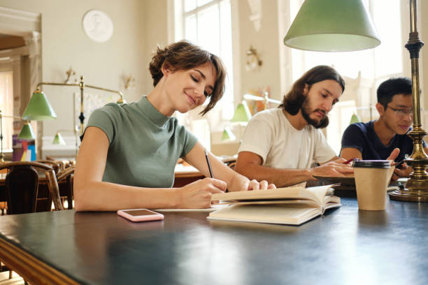 jeune étudiante séduisante avec un livre travaillant joyeusement sur un projet d’étude à la bibliothèque de l’université - book working college student classroom photos et images de collection