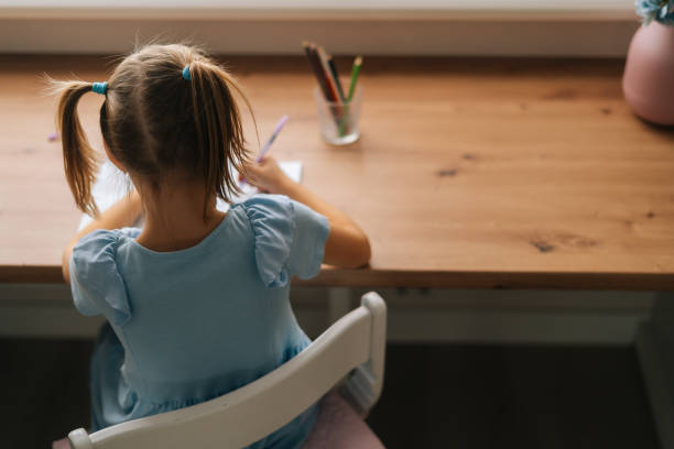 istock High-angle back view of unrecognizable little child girl learning writing doing homework sitting at home table by window. 1358966695