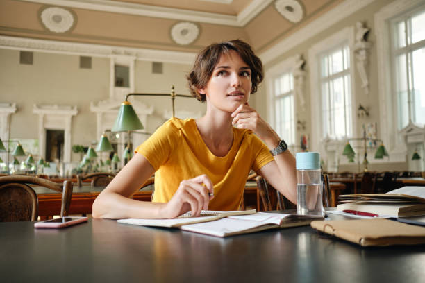 istock Young beautiful casual female student with notepad dreaming while study in library of university 1358967605