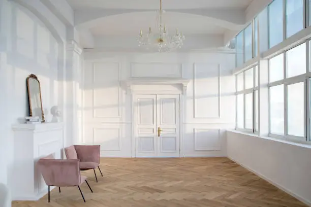 Interior of spacious white hall with armchairs placed near artificial fireplace with statuette and mirror on it opposite wide windows and big chandelier hanging from ceiling