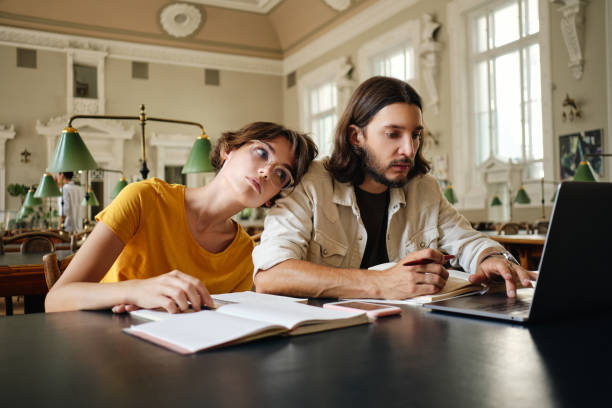 istock Two tired students thoughtfully working on study project with laptop and book in library of university 1358967915