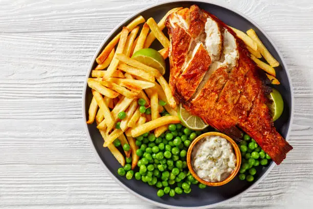 fried Atlantic redfish on a plate with french fries, boiled green peas, tartar sauce and lime slices, horizontal view from above,  flat  lay, close-up, free space