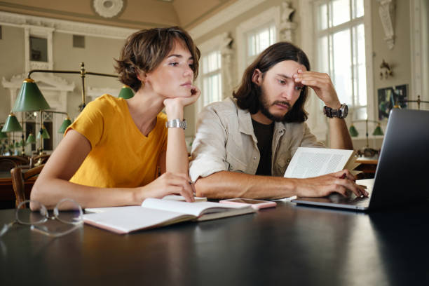 istock Two young pensive students thoughtfully working on study project with laptop and books in library of university 1358968159