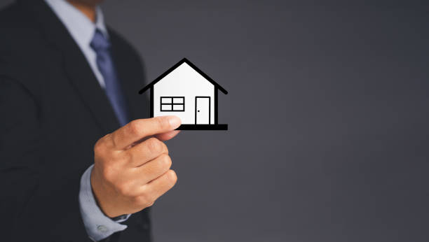 istock A businessman's holding a mini paper house while standing over gray background 1358969147