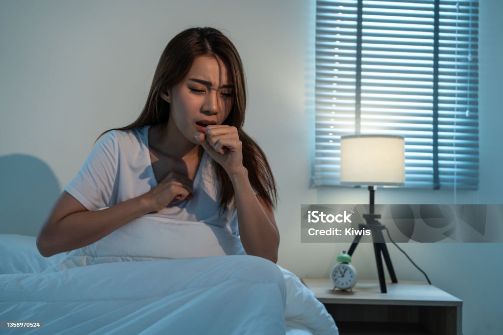 Asian beautiful girl in pajamas feel sick while sit on bed in bedroom. Attractive sleep young woman cover her mouth, feeling bad with fever and coughing after wake up on bed in the morning at home. Coughing Stock Photo