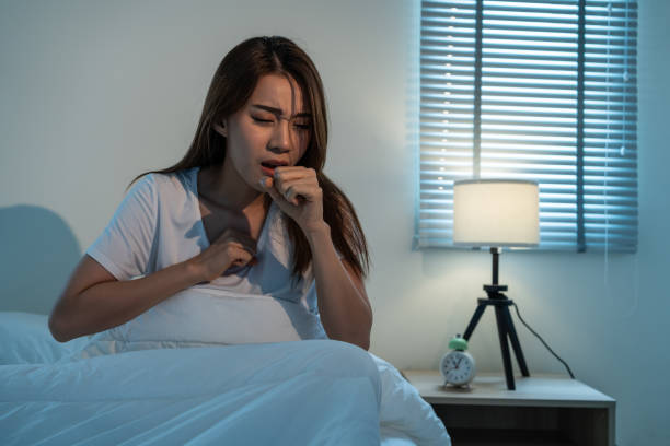 Asian beautiful girl in pajamas feel sick while sit on bed in bedroom. Attractive sleep young woman cover her mouth, feeling bad with fever and coughing after wake up on bed in the morning at home. stock photo