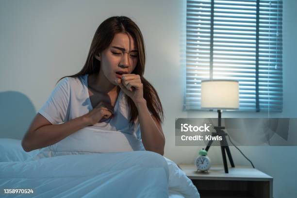 Asian Beautiful Girl In Pajamas Feel Sick While Sit On Bed In Bedroom Attractive Sleep Young Woman Cover Her Mouth Feeling Bad With Fever And Coughing After Wake Up On Bed In The Morning At Home Stock Photo - Download Image Now