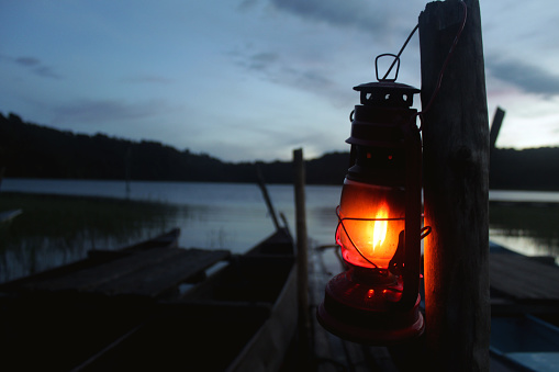 Old classic oil lantern of fisherman hanging on a wood with wooden fishing boats and blue sky over the lake in the early morning.