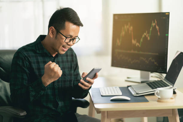 Asian man trader happy and successfully sitting at home office in front of monitors with cryptocurrency graph holding smartphone browsing application monitoring cryptocurrency price comerciante hombre asiático feliz y con éxito sentado en la oficina en casa frente a los monitores con gráfico de criptomonedas que sostiene la aplicación de navegación de teléfonos inteligentes que monitorea el precio de la criptomoneda