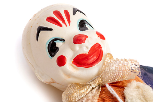 a mad evil redhead clown, wearing a white and red striped costume with a white ruff, stands with his arms on his hips and a creepy smile in front of the weathered tent of the circus