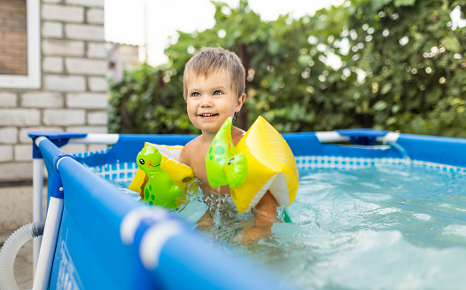 Little naked smiling kid in green swimming trunks with bright inflatable safety oversleeves swims plays and jumps in transparent clean cool water in the pool, in the yard