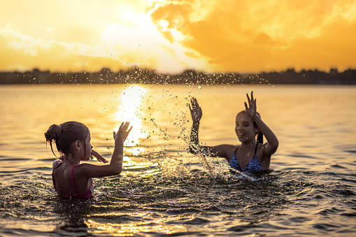 Two funny funny sisters splash on each other with shiny transparent clear water in a blue shallow lake, against the backdrop of a bright sunny golden summer warm sunset