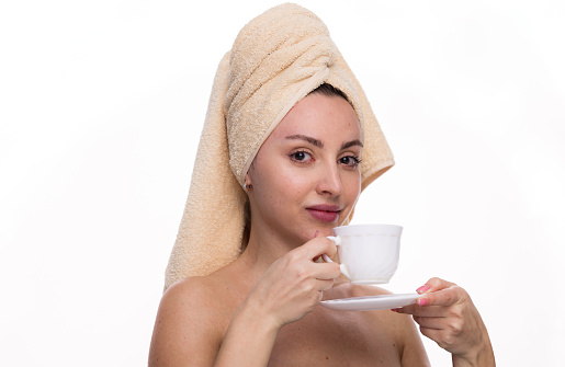 Happy young woman with towel on her head holds cup of coffee or tea in her hand and smiles on white background. Morning routine, nice time spent