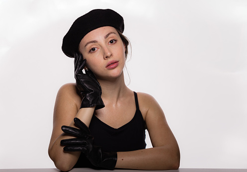 Portrait of gorgeous young fashion model in black beret and leather gloves leaning on the table on white background. Closeup face of young beautiful woman with a healthy clean skin