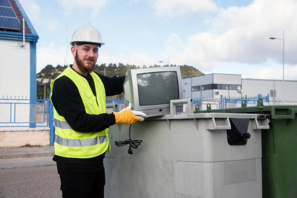 recycling center worker posing next to and old tube television and dumpsters recycling center worker posing next to and old tube television and dumpsters. Man wearing protective gloves and helmet and a reflective vest. Concept of recycle electronics recycling center stock pictures, royalty-free photos & images