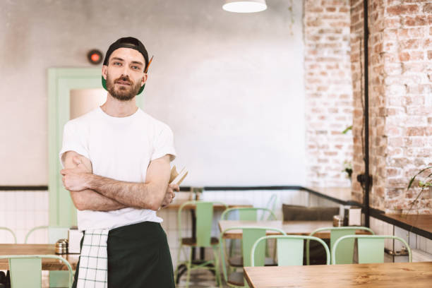 istock Young man in black cap,white t-shirt and apron standing and dreamily looking in camera while working in cafe 1358993694