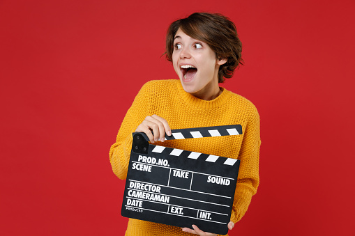 Excited surprised young brunette woman 20s wearing casual yellow sweater standing holding classic black film making clapperboard looking aside isolated on bright red colour background studio portrait