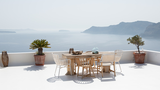 Whitewashed rooftop terrace with trendy wooden chairs at rustic wooden table outdoors. View of Caldera, Cyclades, Greece.