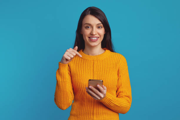 istock Young cute girl pointing at smartphone while smiling against blue background 1358999193