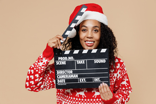 Happy young american Santa woman in Christmas hat sweater holding classic black film making clapperboard isolated on pastel beige background studio portrait. Happy New Year merry x-mas holiday concept