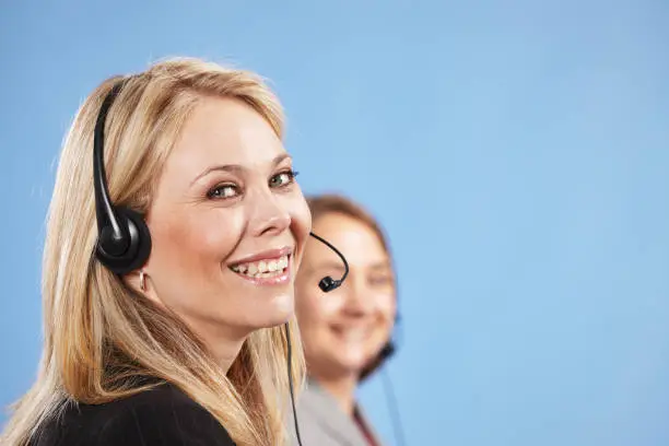 Happy, smiling woman in foreground wearing headphones and microphone, with her colleague in the background.