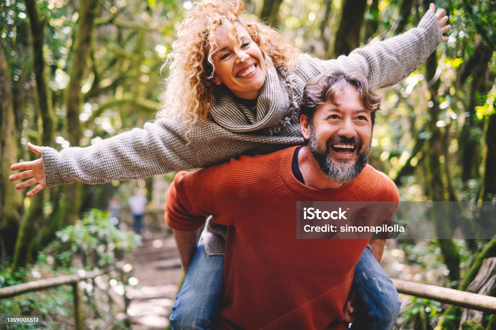 Overjoyed adult couple have fun together at outdoor park in leisure activity. Man carrying woman in piggyback and laugh a lot. Love and life mature people lifestyle concept. Enjoying vacation nature Couple - Relationship Stock Photo