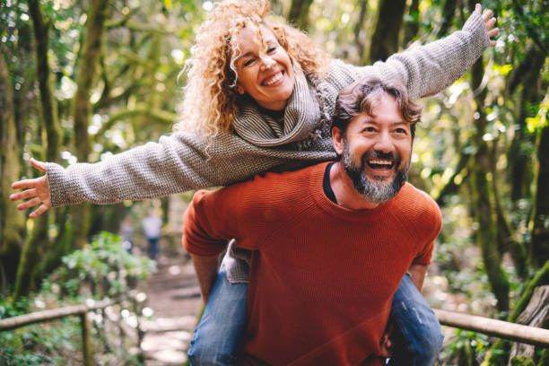Overjoyed adult couple have fun together at outdoor park in leisure activity. Man carrying woman in piggyback and laugh a lot. Love and life mature people lifestyle concept. Enjoying vacation nature stock photo
