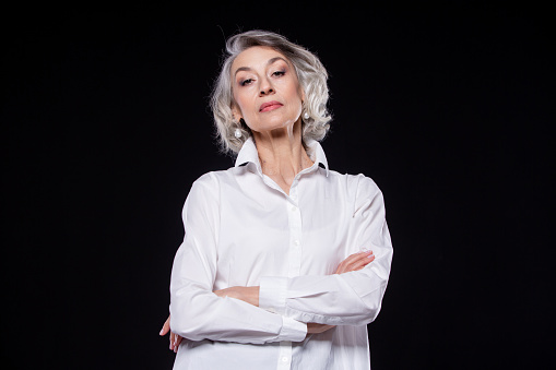 Portrait of a domineering, arrogant mature woman looking down and standing with her arms crossed isolated on a black background.