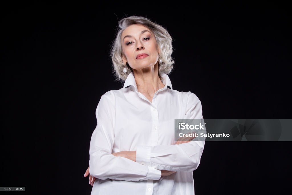 Portrait of a domineering, arrogant mature woman looking down and standing with her arms crossed isolated on a black background Portrait of a domineering, arrogant mature woman looking down and standing with her arms crossed isolated on a black background. Arrogance Stock Photo