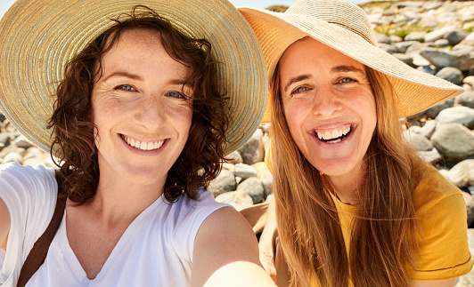 Close-up portrait of smiling female friends in sun hat standing together at the beach taking selfie