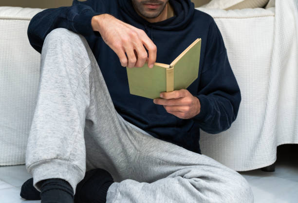 młody mężczyzna w wygodnym ubraniu czytający książkę przy łóżku w domu. przypadkowy koleś czytający książkę w domu. - sitting young men book isolated on white zdjęcia i obrazy z banku zdjęć