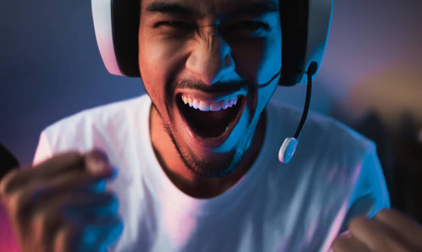 Young Asian man playing online computer video game, colorful lighting broadcast streaming live at home. Ecstatic celebration winning a match. Gamer lifestyle, E-Sport online gaming technology concept stock photo