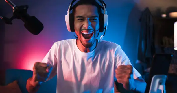 Photo of Young Asian man playing online computer video game, colorful lighting broadcast streaming live at home. Ecstatic celebration winning a match. Gamer lifestyle, E-Sport online gaming technology concept