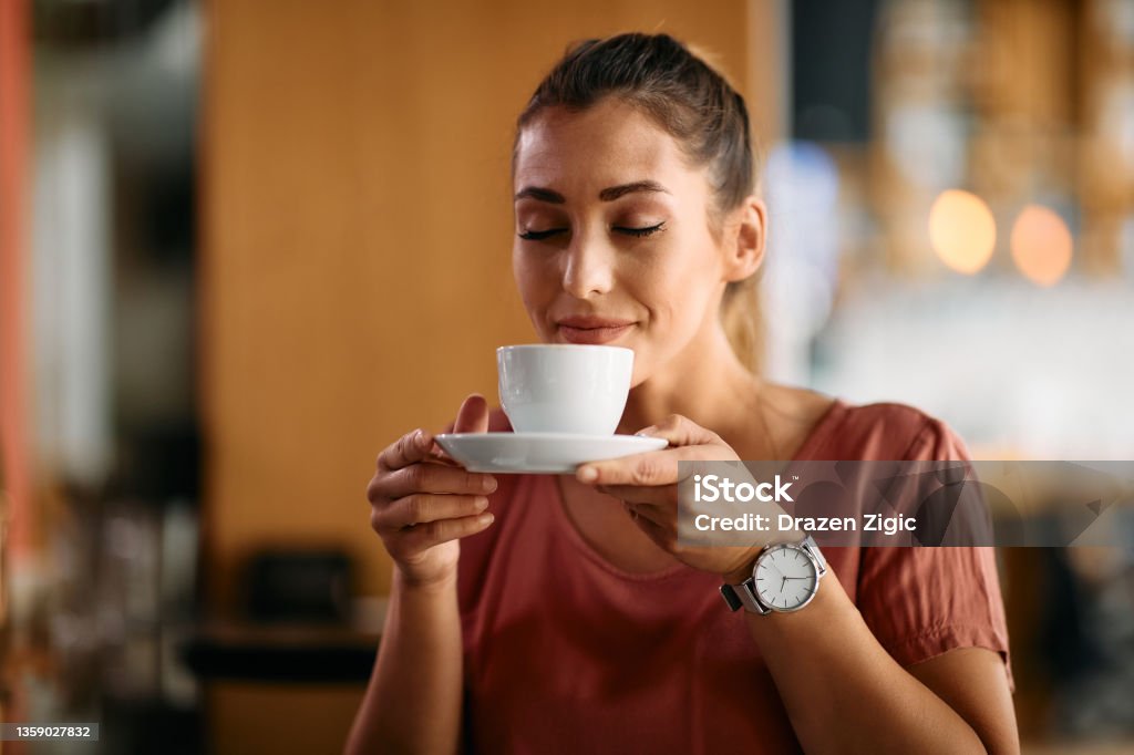 Young woman enjoying in smell of fresh coffee in a cafe. Smiling woman enjoying with eyes closed in cup of fresh coffee at cafe. Coffee - Drink Stock Photo