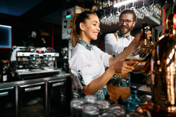 Female bartender pouring beer draft beer while working with colleague in a bar. Young happy bartenders pouring beer from a beer tap while working in a pub. Focus is on woman. bartender stock pictures, royalty-free photos & images