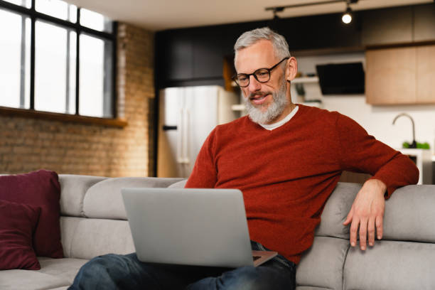 istock Mature middle-aged caucasian man in casual clothes sitting on sofa using laptop for e-banking, e-commerce, distant work, remote education online at home on quarantine 1359037070