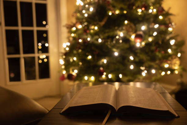 Open bible by Christmas tree Open bible on a wood table in a domestic living room by leather lounge chair and glass door with softly glowing Christmas tree in a dark room Bible stock pictures, royalty-free photos & images