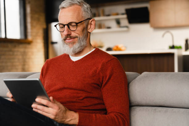 zbliżenie portret uśmiechniętego kaukaskiego dojrzałego mężczyzny w średnim wieku w okularach za pomocą cyfrowego tabletu do e-learningu, płacenia rachunków online, e-commerce, pracy zdalnej, mediów społecznościowych online w domu - senior adult computer men mature adult zdjęcia i obrazy z banku zdjęć