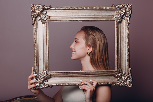 Millenial young woman blonde hair holds gilded picture frame in hands face portrait.