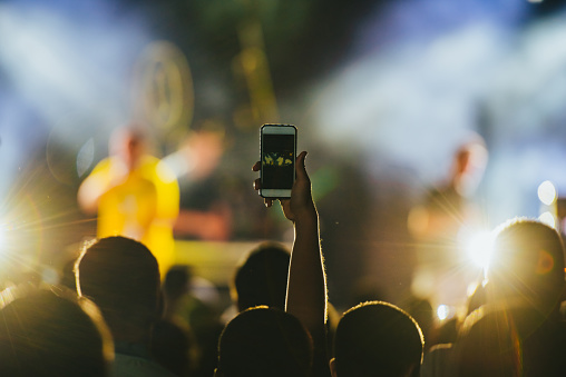 Man hand holding smartphone and taking picture in a concert crowd in front of bright stage lights on a music festival
