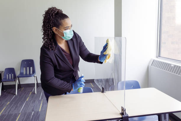 Female teacher cleaning classroom during COVID-19 pandemic A mixed race female teacher wearing a protective face mask and surgical gloves disinfects a desk with a plexiglass divider to protect students from infection during the COVID-19 pandemic. Cleanliness of the school: stock pictures, royalty-free photos & images