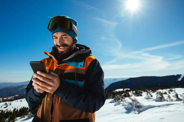 Finding time for his social media Smiling bearded snowboarder typing on his mobile phone while on the mountain skiing photos stock pictures, royalty-free photos & images