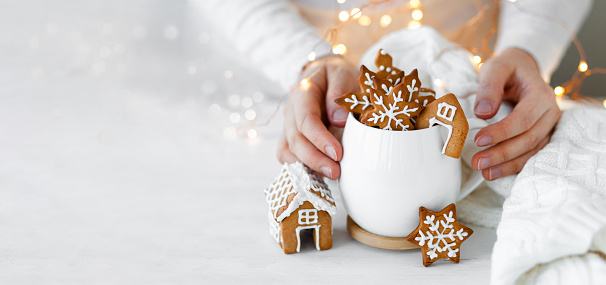 Young woman holding white mug with hot chocolate and gingerbread cookies. Holiday atmopshere, cozy handmade decor. Lights on background, bokeh. Close up copy space for text, banner