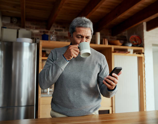 content multi-cultural elderly male taking a sip of his morning coffee while catching up on news on his smartphone, standing in modern kitchen - senior adult human face male action imagens e fotografias de stock