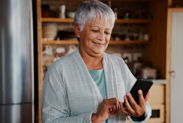 istock Smiling multi-cultural elderly female typing a message on smartphone while standing in modern kitchen 1359088578