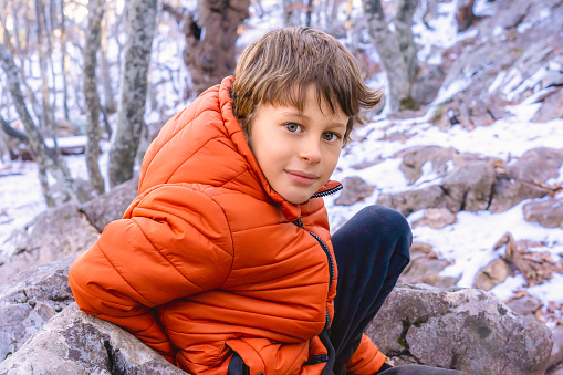 A white Caucasian boy 8 years old in a pink jacket sits in a winter forest in the mountains while resting during a hike