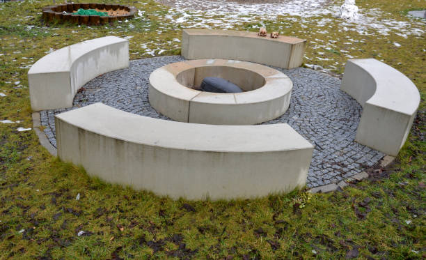 fireplace in a paved stone circle in the garden and park. in the middle is a concrete stone fireplace to ignite the wood. around are symmetrically placed curved rectangle benches and pallisade sandbox chimenea en un círculo de piedra pavimentada en el jardín y el parque. en el medio hay una chimenea de piedra de hormigón para encender la madera. alrededor hay bancos rectangulares curvos colocados simétricamente y caja de arena pallisade