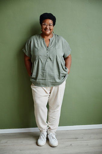 Vertical full length portrait of black senior woman wearing glasses and smiling at camera while standing against green background