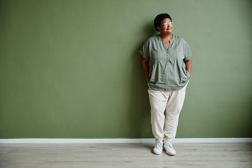 Full length portrait of black senior woman wearing glasses and smiling while standing against green background indoors, copy space